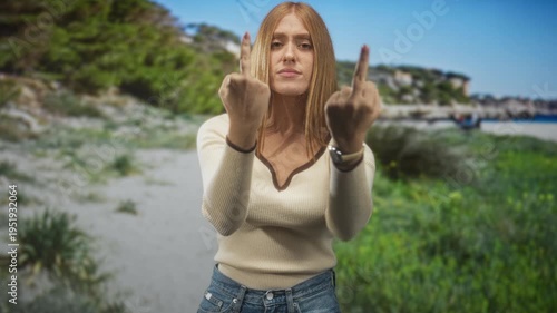 Redhead woman shows both middle fingers to camera with tense hands at a beach shoreline, wearing cream sweater and jeans, stern expression; defiance.
