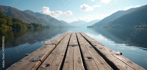 Rustic wooden pier extends onto calm lake water, backed by tree-lined hills and hazy mountains under clear blue sky. Serene natural landscape invites peaceful outdoor activity and quiet contemplation.