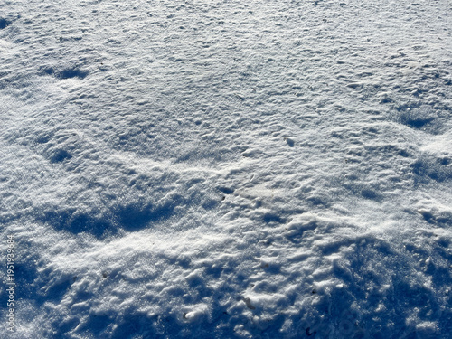 Close‑up of uneven, sunlit snow surface with natural ripples, sparkles, and subtle shadows, creating a bright winter texture with crystalline highlights.