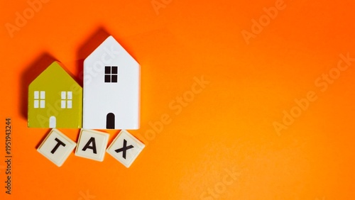 Quadro su tela Two miniature model houses are placed above letter tiles spelling TAX on a vibrant orange surface, symbolizing property taxes, real estate liabilities, and the financial costs of homeownership