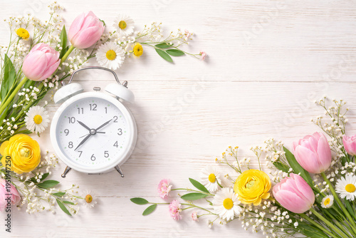 White alarm clock with delicate white blossoms on a light wooden table, soft blue background and copy space, minimal spring time concept with clean aesthetic.