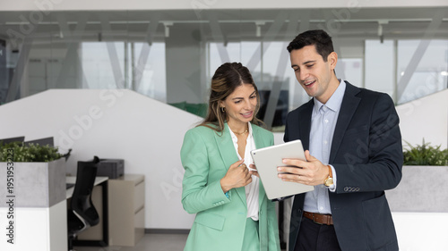 Obraz na plátně Confident young adult business professional mentoring female coworker using tablet in modern office guiding in using digital tools making task review in friendly collaborative atmosphere