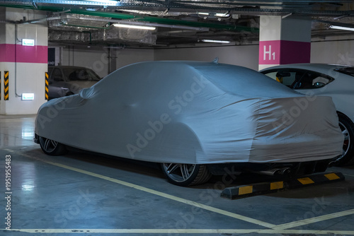 Mysterious luxury car, concealed beneath light grey protective cover, securely parked in dimly lit, modern underground garage with concrete pillars and visible utilities