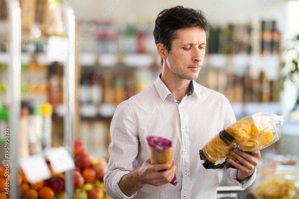 Fototapeta premium Man in light shirt took some varieties of pasta from shelf. Client compare pasta noodles items package. Customer examines contents of package, choose noodles garnish product