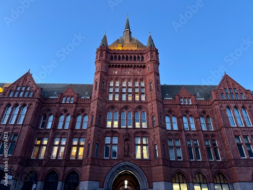 Foto [Intimidating Medieval Brick Grandeur]: A grand, regal, and multi-story red brick building with complex medieval inspiration and thousands of windows, dominating a European street
