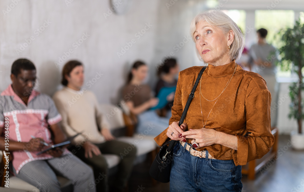 Obraz premium Thoughtful mature woman standing while people waiting in background in reception area