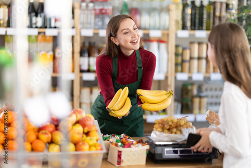 Friendly young saleswoman giving bananas to teenage female customer at grocery shop