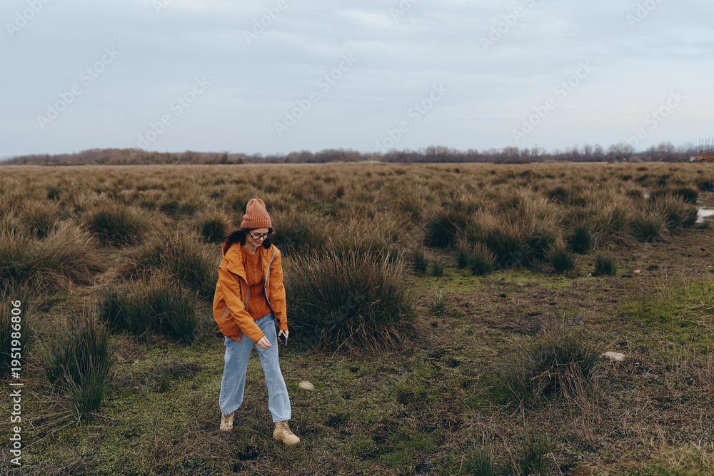 Obraz premium Woman lifestyle concept with a young woman wearing an orange jacket and hat walking in a vast grassy field under an overcast sky during autumn season outdoors.