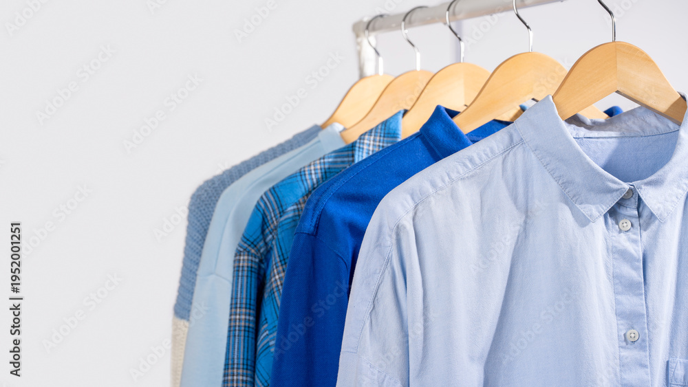 Fototapeta premium Close up blue shirts hanging on wooden beige hangers on clothing rack, light gray background. Concept of capsule wardrobe, minimalist clothing basics and organized closet lifestyle. Copy space, banner