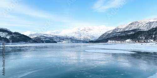 Frozen lake reflects mountains, sky. Winter landscape presents serene beauty, calm waters, snowy peaks. Quiet stillness dominates scene.
