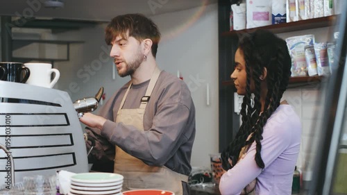 Two multiracial Barista show trainee prepares coffee while colleague observes in a busy cafe during the afternoon shift