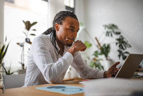 A man with braided hair, wearing a white shirt, sits at a desk with a laptop and a pencil in his mouth, deep in thought.