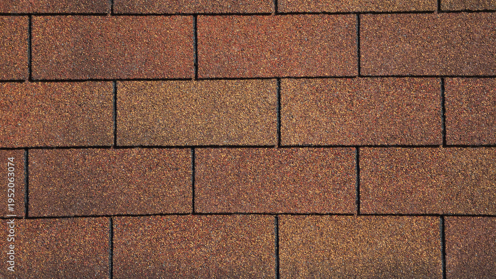 Fototapeta premium A close-up of brown and tan asphalt roof shingles arranged in a staggered rectangular pattern. The surface features a gritty, granular texture with subtle variations in earth-toned shades.