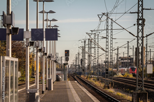 Platform view at Troisdorf train station in Germany, with dense railway wires, signals and a Cologne Koln S-Bahn train visible on the right. The scene highlights commuter rail infrastructure in NRW.