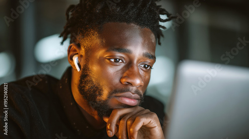 Young black professional man with wireless earbuds concentrating on data displayed on laptop screen in modern coworking office, technology analysis and productivity concept