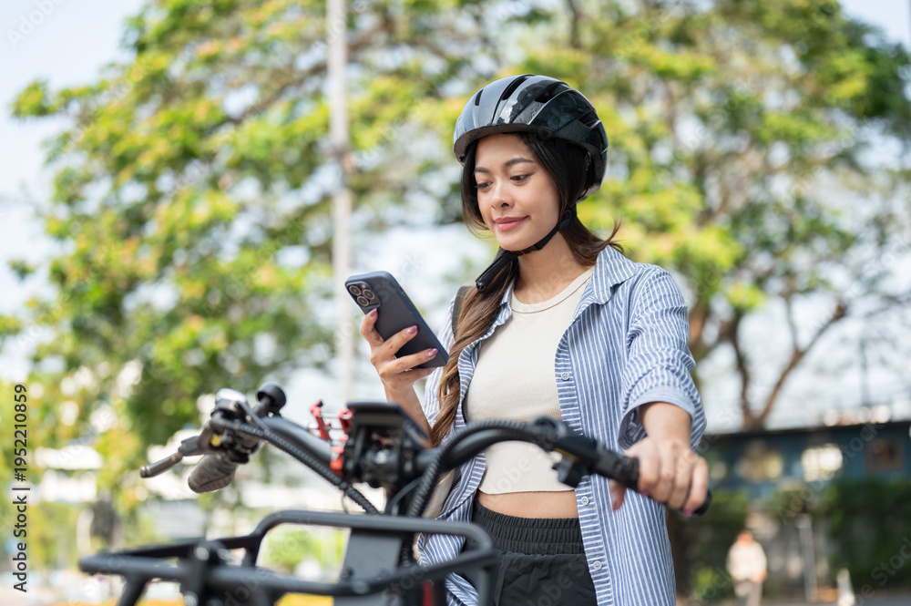 Naklejka premium Pretty asian woman wearing helmet looking at smartphone while riding e bike electric bicycle in park