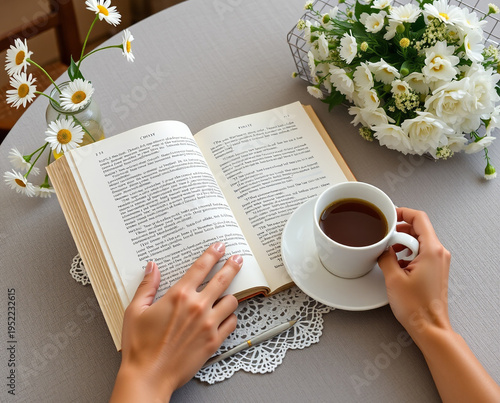 Person reading an open book with a cup of coffee and white daisies on a table