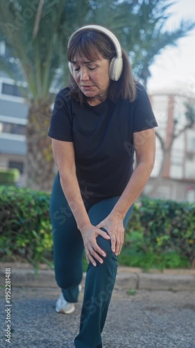 Middleaged woman wearing headphones holds knee while stretching during a street warmup by palm hedge and sidewalk; knee pain concern.