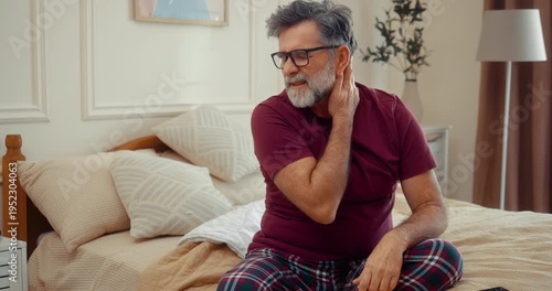 Senior man with neck pain sitting on bed in the morning, feeling stiffness and discomfort