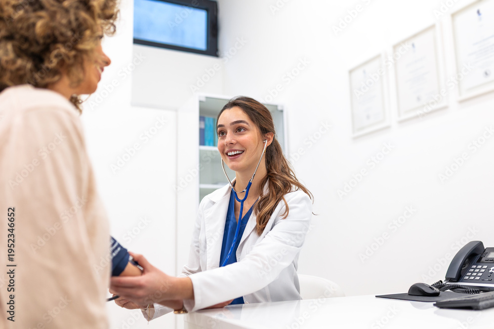 Fototapeta premium Smiling female doctor measuring blood pressure of woman patient in clinic office. Professional practitioner using manual sphygmomanometer. Healthcare and medicine consultation and hypertension concept