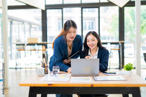 Two colleagues are smiling and working together on laptops at a modern desk.
