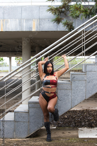 A beautiful, dark-haired Latin American woman dressed as a zombie, wearing a red and black leather top and shorts,black fishnet stockings and black boots.The woman is leaning on the staircase railing.