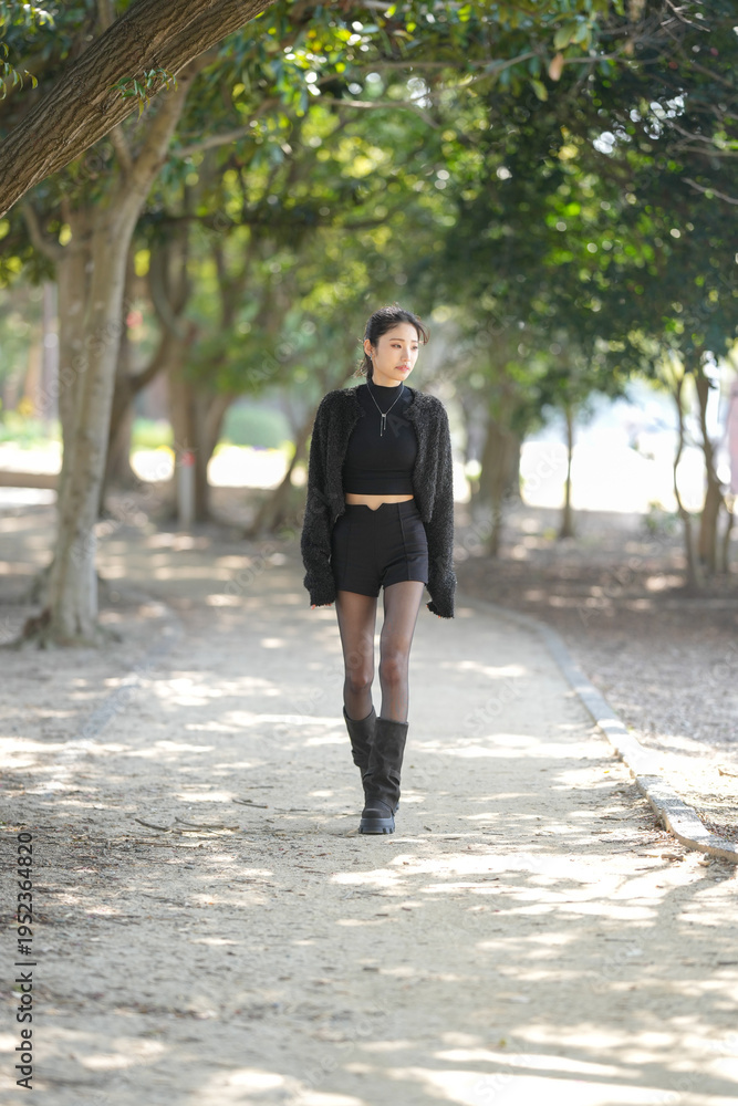 Fototapeta premium Young woman smiles softly, leaning against tree trunk. Black knit top and sparkly cardigan add stylish contrast. Dark shorts and sheer tights complete her trendy look