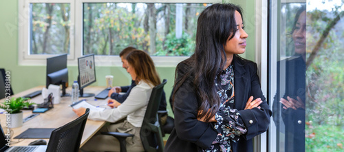 Banner of hispanic young businesswoman with arms crossed gazes out an office window, reflecting on strategy while colleagues work in a modern shared workspace. Copy space available on left.