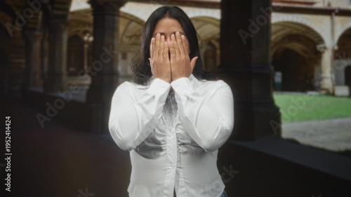 Woman covering eyes with hands in building courtyard wearing white shirt and standing centered in cloister; shyness vulnerability.