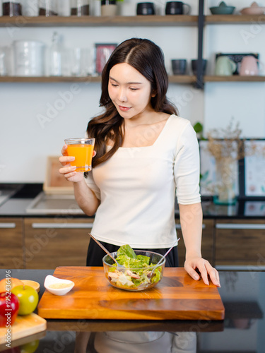 Asian young pretty positive woman standing in kitchen holding drinking orange juice eating healthy salad cooking healthy food fresh vegetables fruits clean feel good morning happy relaxing lifestyle.