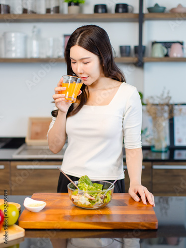Asian young pretty positive woman standing in kitchen holding drinking orange juice eating healthy salad cooking healthy food fresh vegetables fruits clean feel good morning happy relaxing lifestyle.