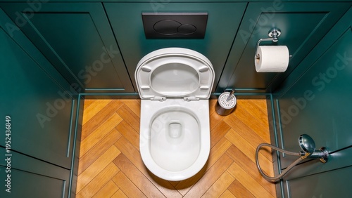 Top view of clean modern toilet with flush plate, toiletpaper holder, brush and bidet sprayer in a stylish bathroom with teal wall and wooden floor.