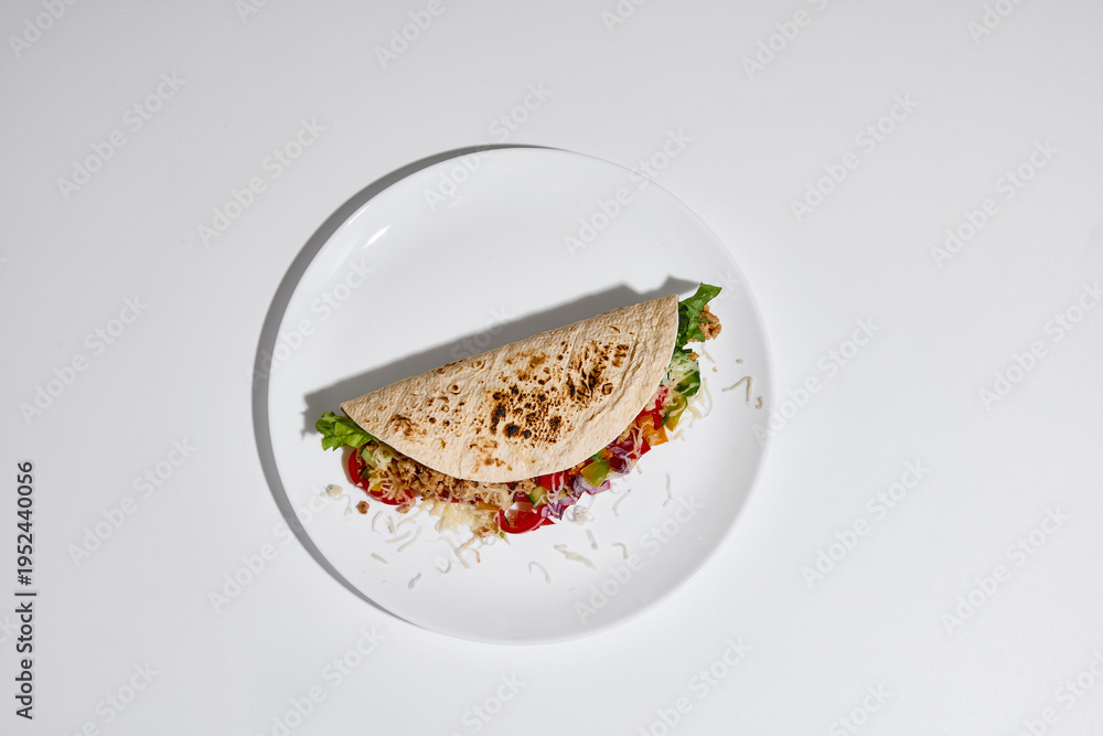 Fototapeta premium Tortilla wrap filled with vegetables, meat and cheese served on white plate on clean white background top view. Concept of street food, takeaway meal, casual dining and food delivery menu design.