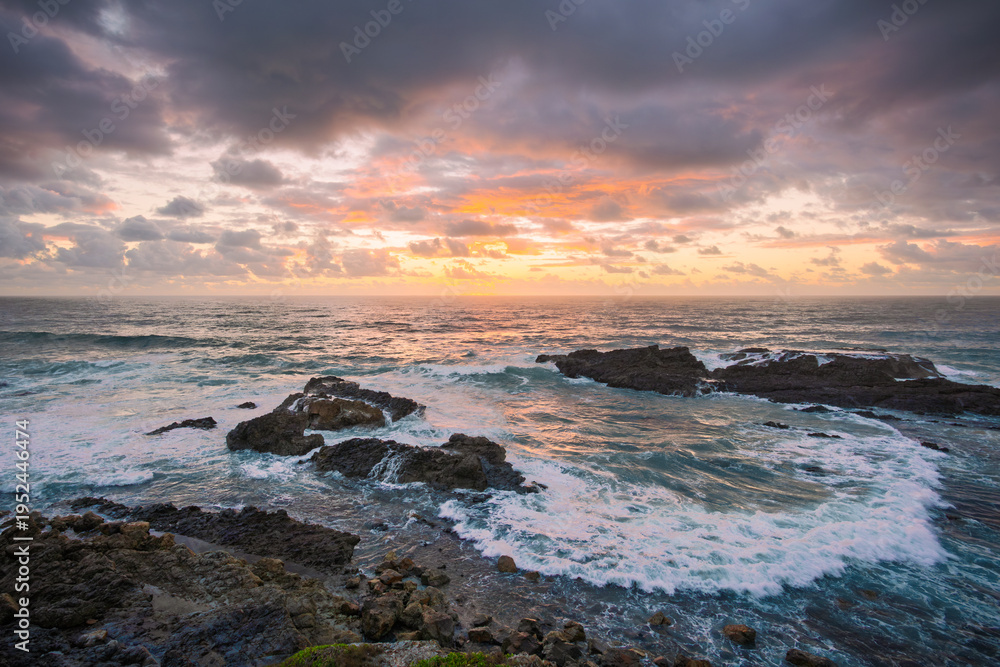 Obraz premium A beautiful sunrise casts an orange glow on the clouds and reflects on the sea near rock outcrops with waves breaking and forming a horseshoe shaped wave at Brooms Head in New South Wales, Australia.