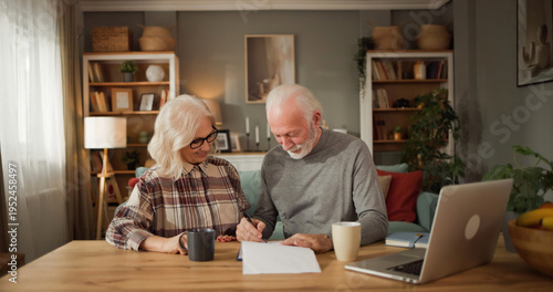 A couple is seated at a table in their home, going over life insurance documents together. The man writes notes while the woman looks on with interest.