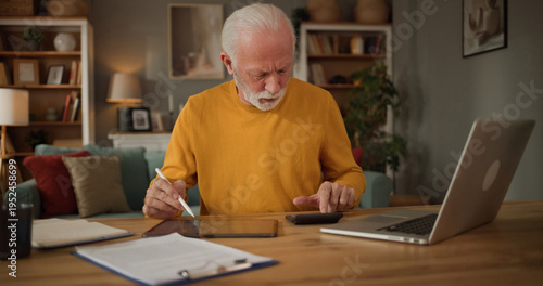 An older man sits at a table in a living room, working on a tablet with a phone nearby. He looks focused as he uses a laptop while organizing his finances in a comfortable space.