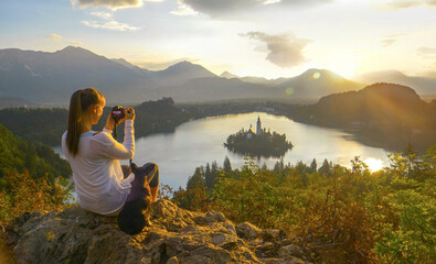 LENS FLARE: Female photographer taking photos of church at lake Bled at sunrise