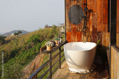 An outdoor bathroom with a serene mountain view. Featuring a standalone bathtub, a rustic wooden wall, and a metallic shower fixture, this space is designed for relaxation and luxury.