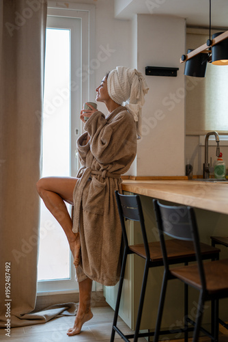 Smiling woman in soft bathrobe with towel on head holding coffee mug in modern kitchen, cozy morning atmosphere, natural daylight, relaxed home lifestyle, wellness and self care concept