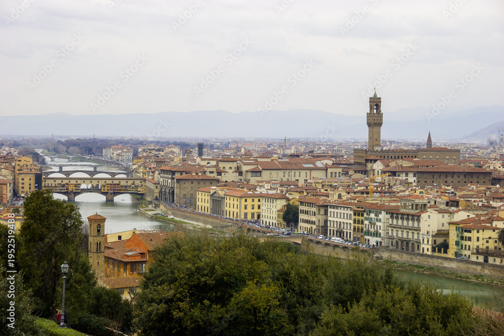 Fototapeta premium A panoramic view of Florence, Italy, showcasing the Arno River, Ponte Vecchio, and the Palazzo Vecchio tower under a cloudy sky.