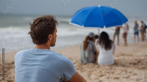 Man in foreground looks towards a blurred beach scene with people under a blue umbrella