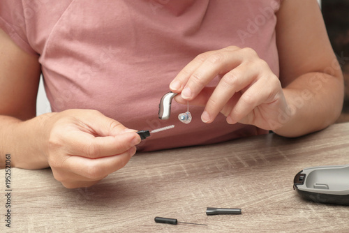 Woman cleaning a digital hearing aid with a small brush tool on a wooden table, hearing aid maintenance and care
