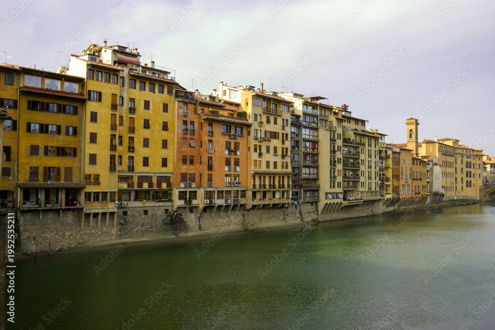 Obraz premium The Arno River flows past the colorful buildings of Florence, Italy, with a bell tower visible in the distance.