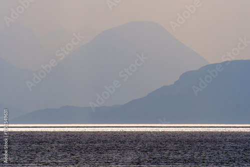 A serene landscape featuring layered, misty mountains fading into the distance, with a shimmering sunlit path across the dark water in the foreground.