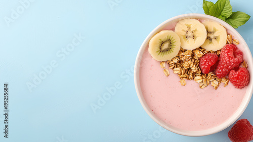 Colorful smoothie bowl topped with sliced banana, kiwi, fresh raspberries, and granola on light blue background, creating fresh and healthy breakfast option