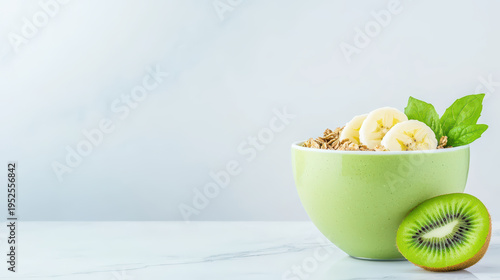 Green bowl filled with granola, banana slices, and fresh spinach leaves next to halved kiwi fruit on light marble surface, evoking fresh and healthy breakfast mood