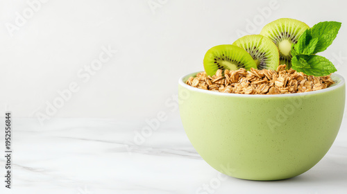 Green bowl filled with granola topped with fresh kiwi slices and mint leaves on white marble surface, offering healthy and refreshing breakfast option