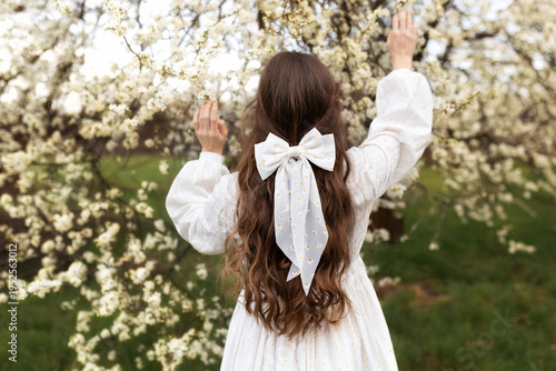 Wallpaper Mural Back view of a young woman with long hair and a white bow, standing in a blooming orchard and reaching toward delicate spring blossoms. Concept of spring, beauty, femininity, and harmony with nature. Torontodigital.ca