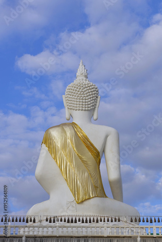 A serene back view of a magnificent white Buddha statue adorned with a shimmering golden robe, sitting under a bright blue sky with soft white clouds.