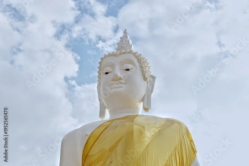 A majestic low angle shot of a serene white Buddha statue adorned with a shimmering golden robe, set against a bright sky filled with soft white clouds.